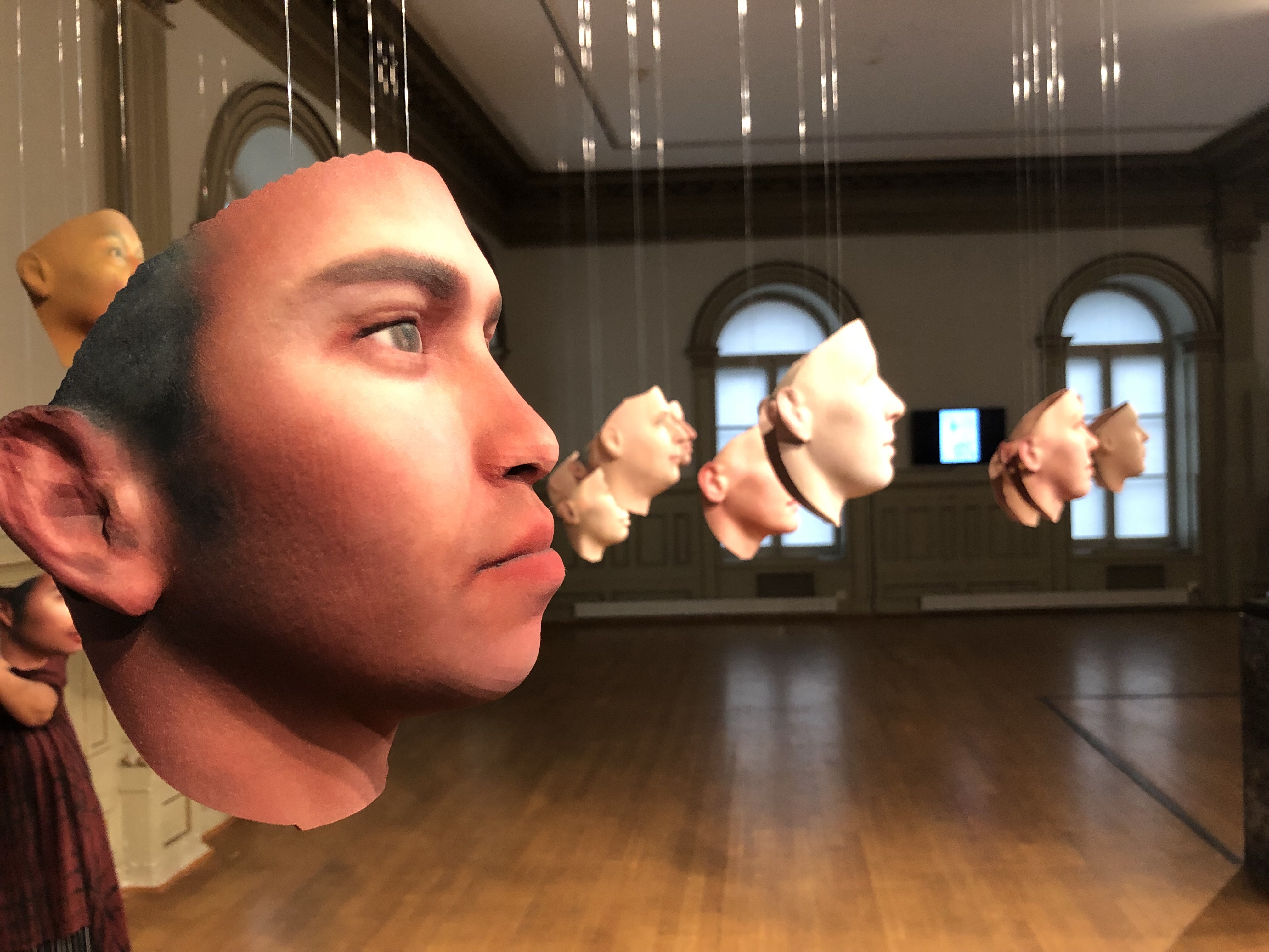 A mask of a photorealistic human face is hanging in the foreground. Other masks hang behind it. 